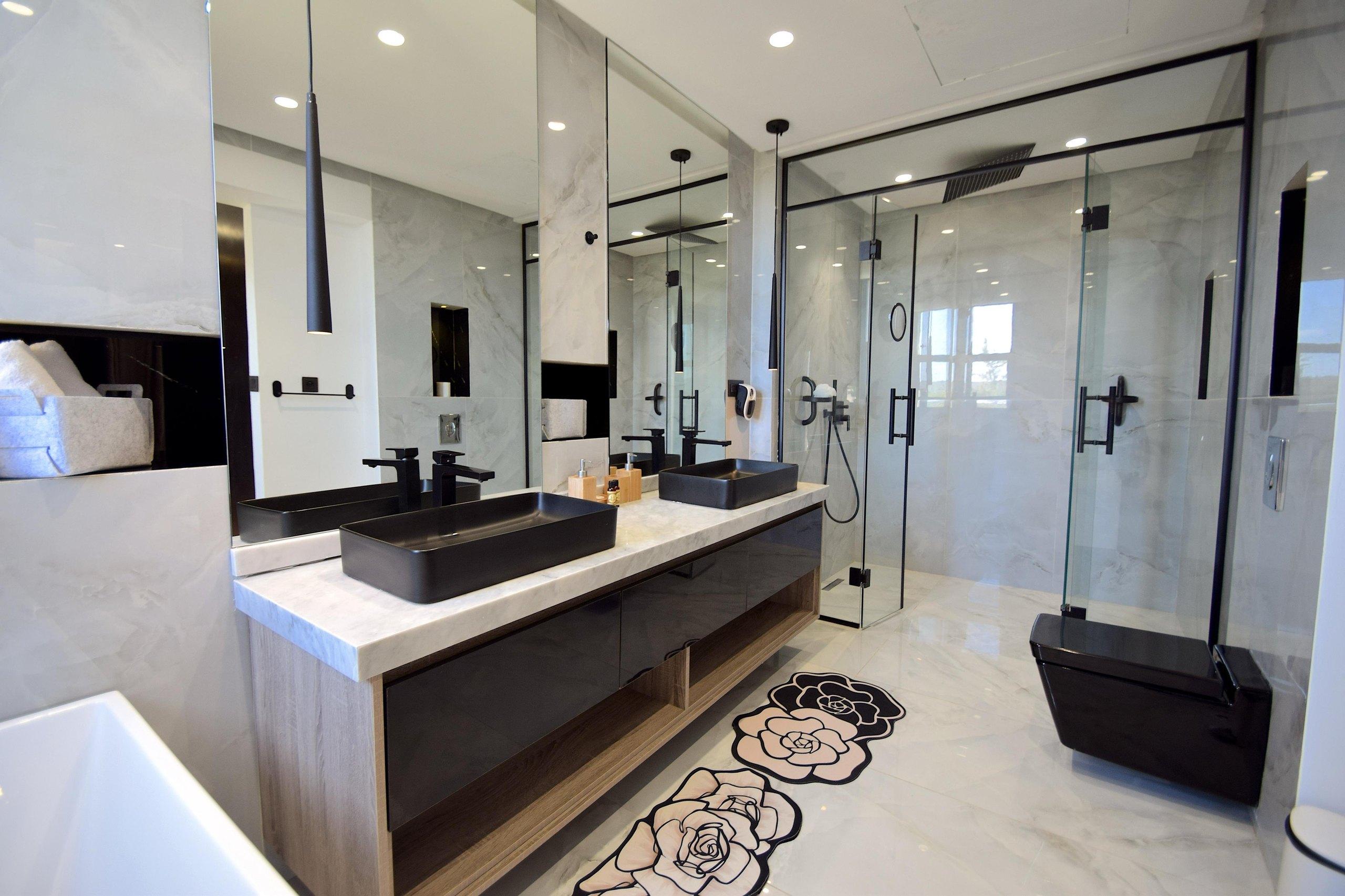 Salle de bain en marbre blanc, deux vasques noires sur plan de marbre et grande douche vitrée à cadre noir.