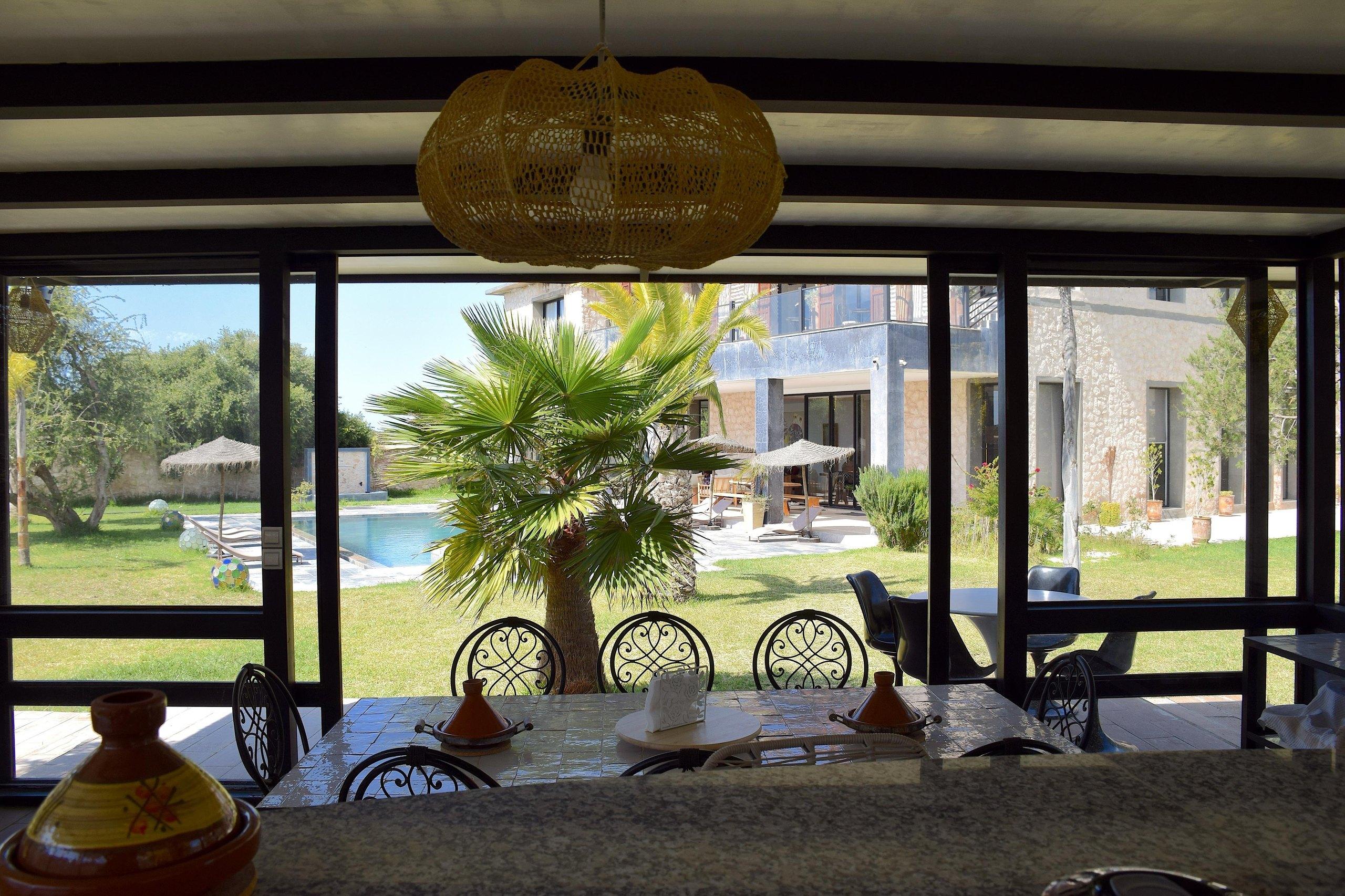 La cuisine extérieure sous son auvent : comptoir de granit, tables en fer forgé et la piscine au-delà des baies.