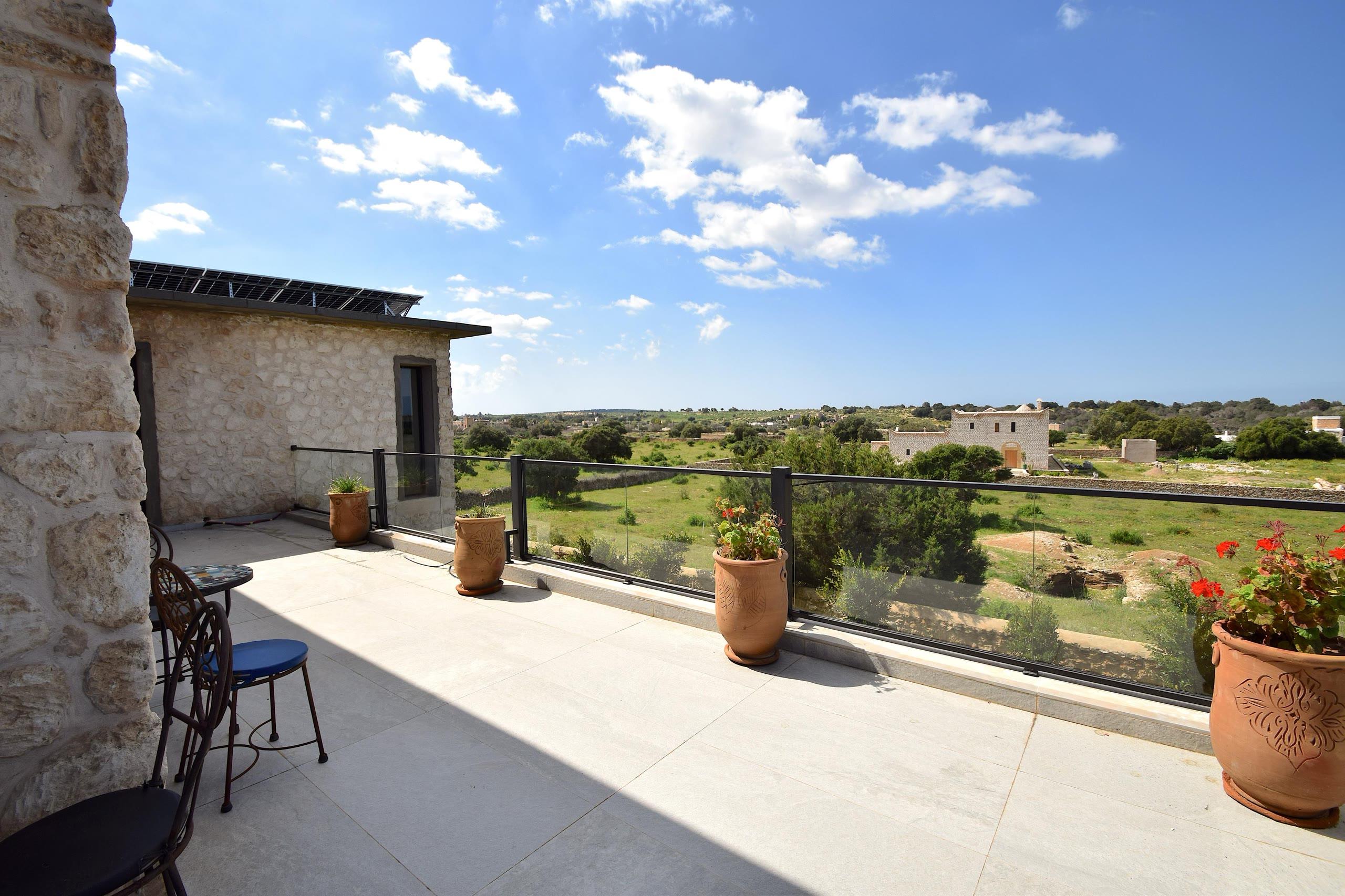 Une terrasse d'étage en pierre claire, garde-corps de verre, ouverte sur les arganiers et la campagne d'Essaouira.