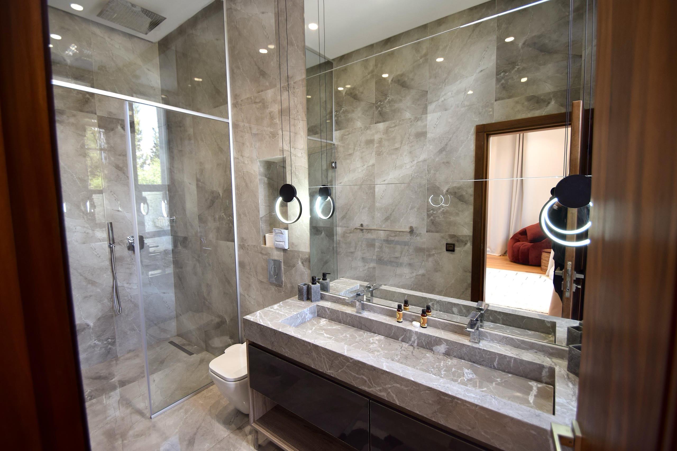 Salle de bain en marbre gris fumé : vasque taillée dans la masse, douche vitrée et porte ouverte sur la chambre.