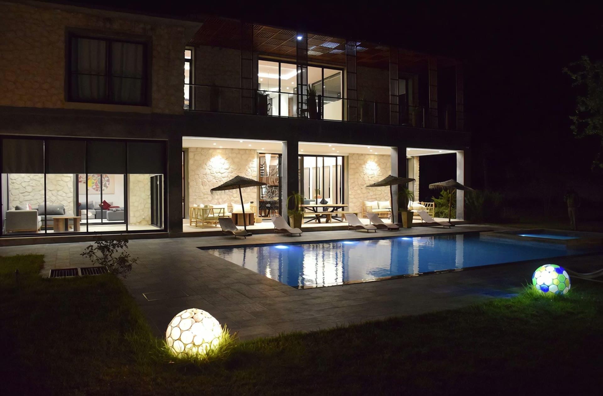 La villa éclairée de l'intérieur à la nuit tombée ; la piscine reflète les baies vitrées.